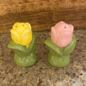 May include: A pair of ceramic salt and pepper shakers shaped like tulips. One is yellow, the other is pink, both with green leaves. The shakers are designed to resemble blooming flowers.