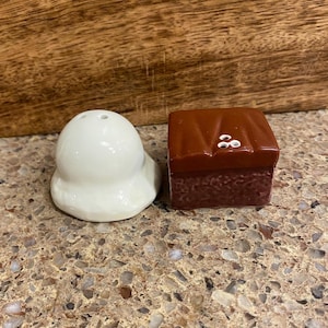 Könnte beinhalten: Ein weißer Salzstreuer aus Keramik und ein brauner Keramikwürfelzucker mit weißen Akzenten. Der Salzstreuer hat eine abgerundete Form mit ausgestelltem Boden. Der Würfelzucker hat eine quadratische Form mit strukturierter Oberfläche.