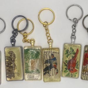 以下が含まれることがあります： ヴィンテージ切手デザインの長方形キーホルダー6個セット。各キーホルダーには、魚、象、鳥など、異なる画像が使用されています。シルバー、ブラック、ゴールドトーンのキーリングとチェーンが付いています。