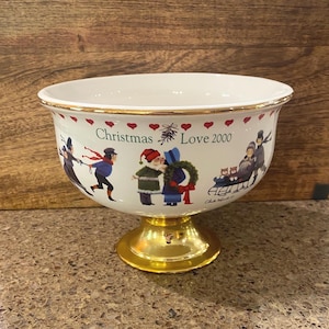 May include: A white ceramic Christmas bowl with a gold-colored base and rim. The bowl features a festive design with figures, hearts, and the text "Christmas Love 2000". The rim is decorated with red hearts.
