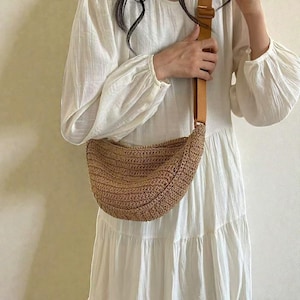 May include: A crescent-shaped shoulder bag in a light brown colour, made from woven material. The bag has a textured, crocheted design and a tan-coloured adjustable strap. The bag is worn over the shoulder, with a leather-like strap.