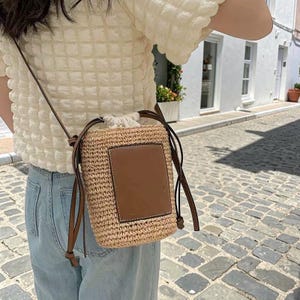 Puede incluir: Un bolso cruzado de paja tejida con un parche de cuero marrón y cierre de cordón. El bolso tiene una correa larga y delgada de color marrón. El bolso es rectangular y parece ser un accesorio de verano.