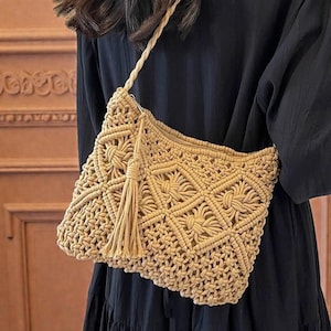 May include: A beige macrame crossbody bag with a woven geometric pattern and a tassel. The bag has a long strap and is worn over a black dress. The bag is made of a natural fibre.