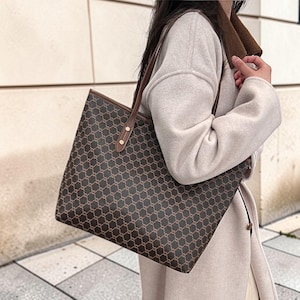 Puede incluir: Un bolso tote grande con exterior marrón oscuro y un patrón geométrico repetitivo en un tono bronceado más claro. El bolso incluye correas de aspecto de cuero marrón sujetas con remaches de metal dorado en el borde superior.