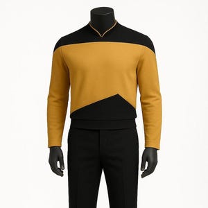 Könnte beinhalten: Eine senfgelbe und schwarze Uniform, bestehend aus einem langärmeligen Oberteil und einer schwarzen Hose, wird auf einer Schaufensterpuppe präsentiert. Das Oberteil hat einen schwarzen Einsatz und ein markantes geometrisches Design. Die Hose ist gerade geschnitten und schwarz.