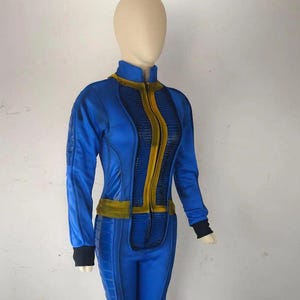 Puede incluir: Mono azul con detalles dorados, cuello alto y cremallera completa. El disfraz tiene una silueta ajustada con paneles texturizados y puños negros. El atuendo se exhibe en un maniquí.