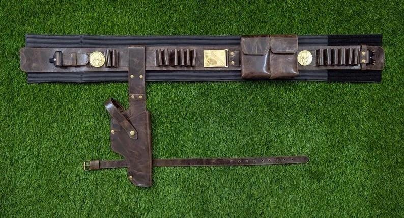 May include: A brown leather gun belt with a holster, pouches, and cartridge holders. The belt has a black elastic backing and gold-coloured accents. The belt is lying on a green surface.