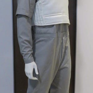 Pu&ograve; includere: Un manichino indossa una tuta grigia e un gilet bianco. La tuta ha maniche lunghe e un collo alto. Il manichino &egrave; in piedi su un disco di metallo.