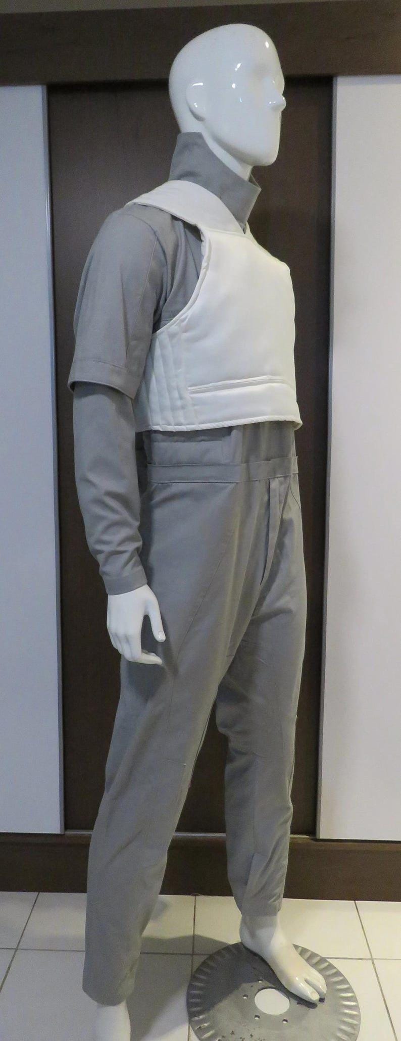 Pu&ograve; includere: Un manichino indossa una tuta grigia e un gilet bianco. La tuta ha maniche lunghe e un collo alto. Il manichino &egrave; in piedi su un disco di metallo.