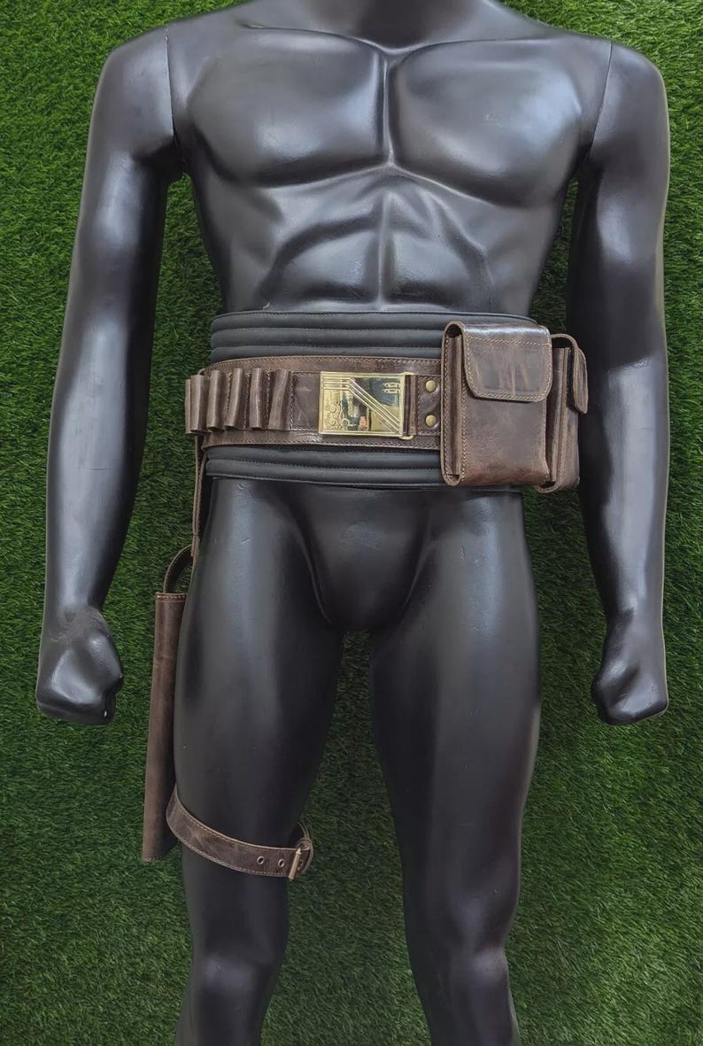 May include: A brown leather belt with a gold buckle and a matching pouch and holster. The belt has multiple loops for holding items. A leg strap is also included. The mannequin is standing on a green surface.