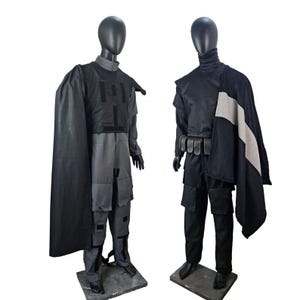 May include: Two mannequins display futuristic-style costumes. The first mannequin wears a gray and black outfit with a cape. The second mannequin wears a black outfit with a cape and a belt with pouches. Both outfits have a tactical appearance.