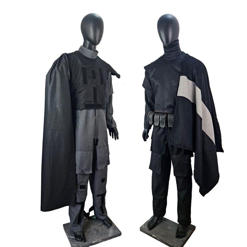 May include: Two mannequins display futuristic-style costumes. The first mannequin wears a gray and black outfit with a cape. The second mannequin wears a black outfit with a cape and a belt with pouches. Both outfits have a tactical appearance.