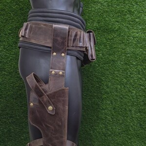 May include: Brown leather holster and belt set. The holster is attached to a leg strap and the belt has multiple pouches. The leather has a distressed look with brass-coloured rivets. The items are displayed on a black mannequin against a green background.