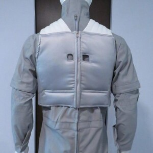 Pu&ograve; includere: Tuta grigio chiaro con gilet abbinato, esposta su un manichino. Il gilet ha una chiusura a zip e due ritagli quadrati. La tuta ha maniche lunghe e un collo alto. Design elegante e futuristico.