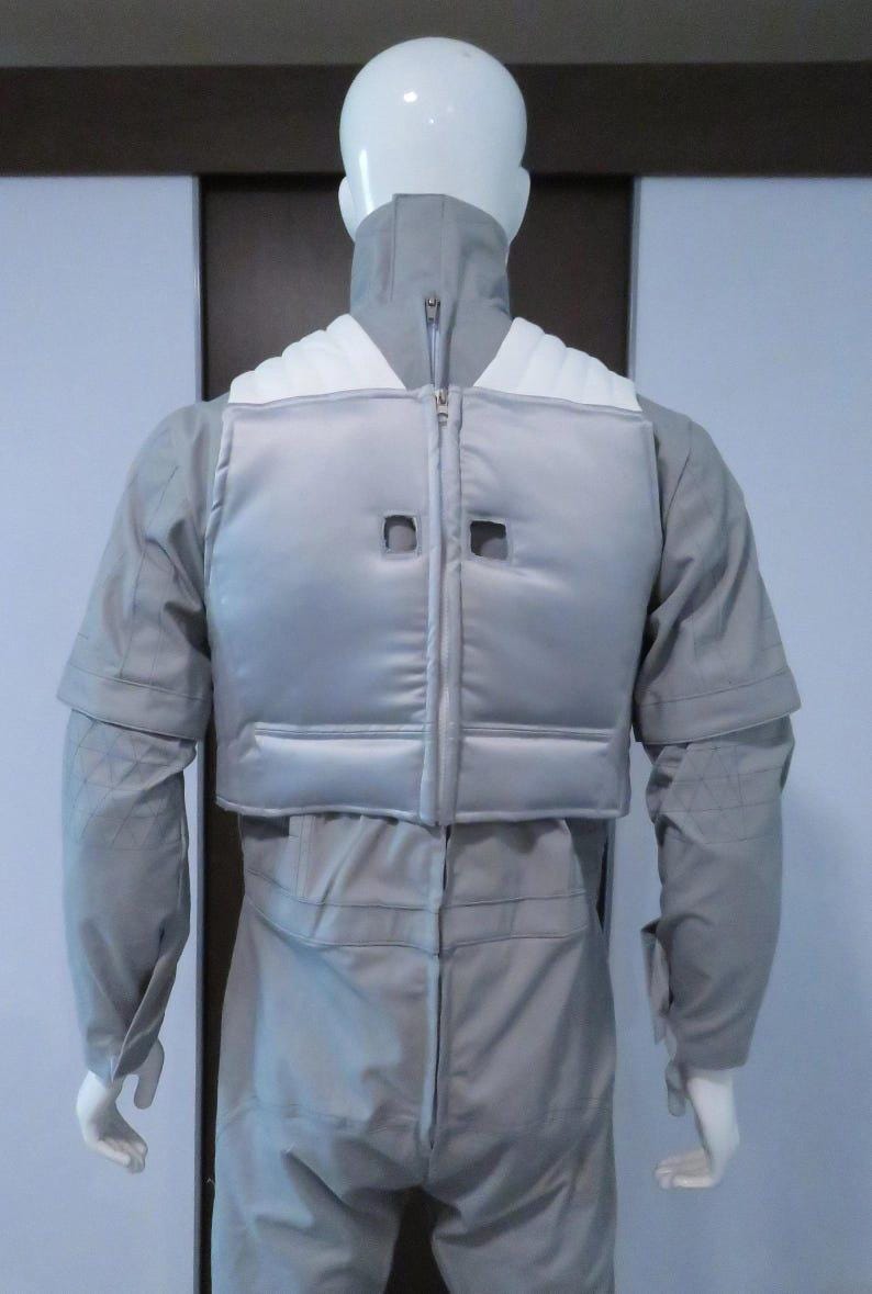 Pu&ograve; includere: Tuta grigio chiaro con gilet abbinato, esposta su un manichino. Il gilet ha una chiusura a zip e due ritagli quadrati. La tuta ha maniche lunghe e un collo alto. Design elegante e futuristico.