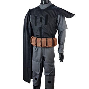 May include: A gray and black costume displayed on a mannequin. The costume includes a gray jumpsuit, a black vest with vertical black stripes, a brown belt with pouches, and a black cape. The mannequin is wearing black gloves and shoes.