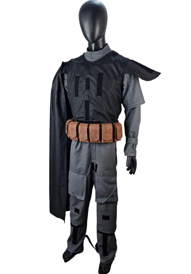 May include: A gray and black costume displayed on a mannequin. The costume includes a gray jumpsuit, a black vest with vertical black stripes, a brown belt with pouches, and a black cape. The mannequin is wearing black gloves and shoes.
