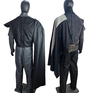 May include: Two mannequins display a dark gray costume with a black cape and a wide belt. The costume includes a long-sleeved shirt, pants, and a high-collared jacket. The cape is draped over the shoulder and back.