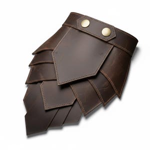 以下が含まれることがあります： 茶色の革製アームガード。鱗のような多層デザイン。真鍮色のボタン留めストラップ付き。革には木目とステッチが見え、手作りのアイテムを示唆しています。