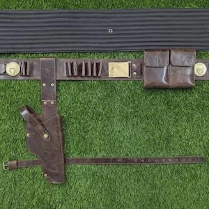May include: A brown leather belt with multiple pouches, cartridge holders, and a holster. The belt has gold-coloured accents and a separate black padded section. A matching brown leather strap is also included. The items are displayed on a green surface.