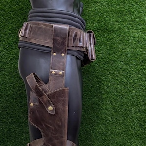 May include: A brown leather holster and belt set. The holster is attached to a leg strap and the belt has multiple pouches. The set is displayed on a black mannequin against a green background.