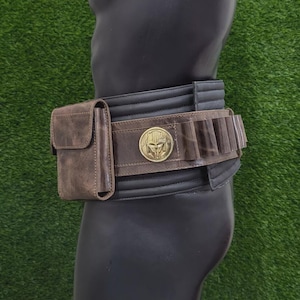 May include: A brown leather belt with a pouch and cartridge holders. The belt has a gold-coloured buckle with a helmet design. The belt is worn on a black mannequin against a green background.