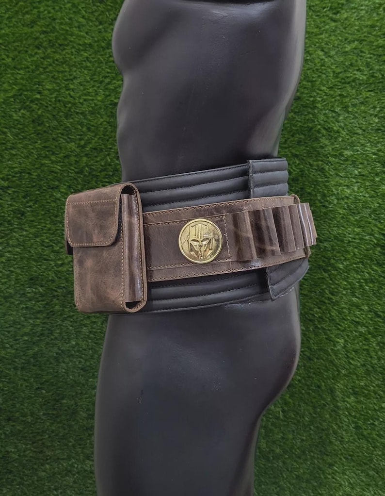 May include: A brown leather belt with a pouch and cartridge holders. The belt has a gold-coloured buckle with a helmet design. The belt is worn on a black mannequin against a green background.