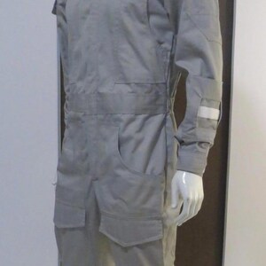 Pu&ograve; includere: Tuta grigio chiaro esposta su un manichino. L'indumento intero presenta un collo alto, maniche lunghe, tasche multiple e un dettaglio in vita. Una toppa circolare &egrave; visibile sulla parte superiore del braccio. Il manichino &egrave; su un pavimento piastrellato.