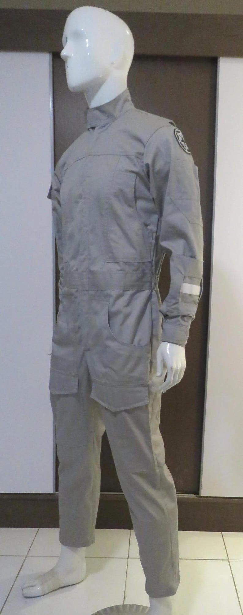 Pu&ograve; includere: Tuta grigio chiaro esposta su un manichino. L'indumento intero presenta un collo alto, maniche lunghe, tasche multiple e un dettaglio in vita. Una toppa circolare &egrave; visibile sulla parte superiore del braccio. Il manichino &egrave; su un pavimento piastrellato.