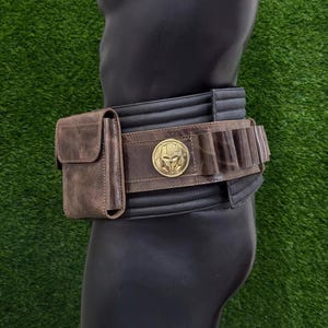 May include: A brown leather belt with a gold emblem and a matching pouch. The belt has a black section with vertical stitching and a brown section with cartridge holders. The belt is displayed on a black mannequin.