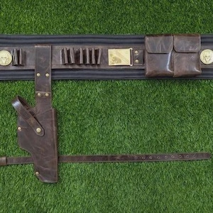 May include: A brown leather gun belt with a holster, pouches, and cartridge holders. The belt has a black backing and is adorned with gold-coloured medallions and a rectangular buckle. The belt is set against a green background.