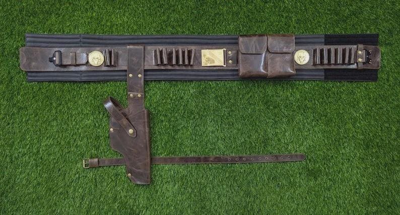 May include: A brown leather gun belt with a holster, pouches, and cartridge holders. The belt has a black backing and is adorned with gold-coloured medallions and a rectangular buckle. The belt is set against a green background.