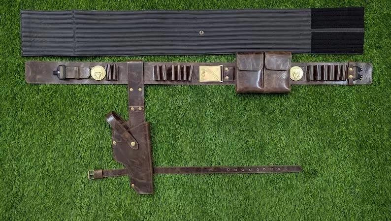May include: A brown leather belt with multiple pouches, cartridge holders, and a holster. The belt has gold-coloured accents and is accompanied by a black, padded support. The items are displayed on a green surface.