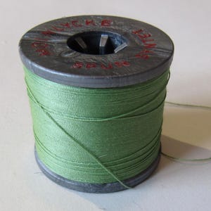 Könnte beinhalten: Eine Spule grünen Fadens, eng um einen grauen Pappkern gewickelt. Der Faden hat einen leuchtenden Grünton, und ein Faden erstreckt sich von der Spule. Die Spule liegt auf einer hellen Oberfläche.