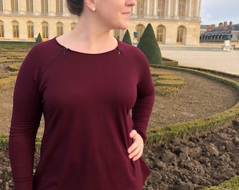 May include: A long-sleeved burgundy top with a round neckline and a slight asymmetrical hemline. The top is paired with blue jeans. The background features a large building with many windows and a manicured garden.