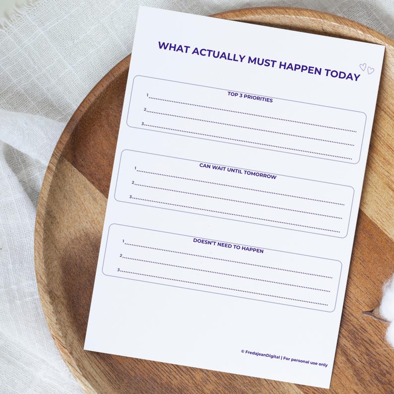 May include: A white paper planner with the text "WHAT ACTUALLY MUST HAPPEN TODAY" in purple. The planner has sections for top priorities, tasks that can wait, and tasks that don't need to happen. The planner is on a wooden tray.