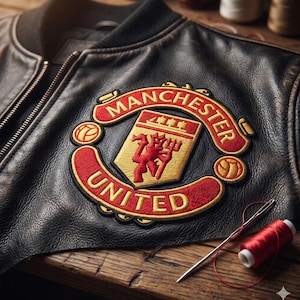 Puede incluir: Chaqueta bomber de cuero negro con el parche del logo del Manchester United. El parche presenta un diablo rojo sobre un escudo dorado, con las palabras "Manchester United" en rojo. Una aguja e hilo rojo sobre una superficie de madera.