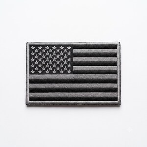 May include: A rectangular embroidered patch featuring a monochromatic American flag design. The patch displays a field of stars on the left and horizontal stripes on the right, all rendered in shades of black, dark grey, and light silver-grey thread.