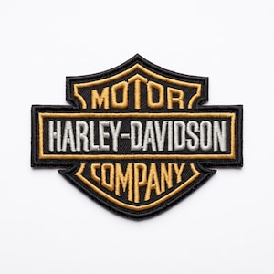 Op de afbeelding: Een schildvormige geborduurde patch met een zwarte achtergrond, gouden randen en belettering. De tekst luidt MOTOR in goud, HARLEY-DAVIDSON in wit en COMPANY in goud. Het ontwerp is een klassiek embleem met een balk en een schild.