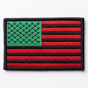 Könnte beinhalten: Ein rechteckiger gestickter Aufnäher mit einem Flaggendesign aus abwechselnden horizontalen roten und schwarzen Streifen. Das Obereck ist ein grünes Quadrat mit einem Muster aus kleinen, erhabenen, schuppenartigen Stickereien.