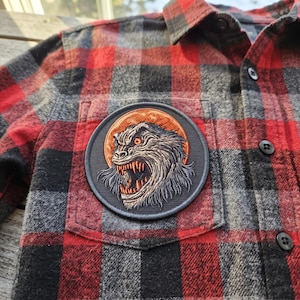 Op de afbeelding: Een overhemd met lange mouwen en knoopsluiting in een rood, zwart en grijs ruitpatroon. Op de borstzak is een ronde geborduurde patch genaaid, met een afbeelding van een grommend, harig wezen met scherpe tanden tegen een oranje achtergrond.