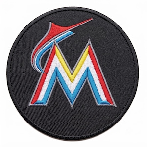 May include: A circular embroidered patch featuring a black background with a stylised letter M in the centre. The letter M is outlined in grey and filled with vertical stripes of red, white, yellow, and blue. A curved red and blue fin shape sits above.