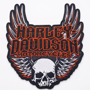 May include: A large embroidered patch featuring the text HARLEY-DAVIDSON MOTORCYCLES in orange lettering on a black background. The design includes white and orange stylised wings extending upwards and a white skull graphic positioned at the bottom.