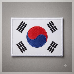 May include: A rectangular embroidered patch featuring the South Korean flag design. The white background displays a central red and blue taegeuk symbol surrounded by four black trigrams positioned in each corner of the fabric patch.