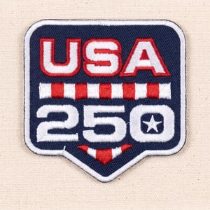 Puede incluir: Un parche bordado con forma de escudo sobre un fondo azul oscuro. El texto USA aparece en blanco con bordes rojos sobre una barra horizontal de rayas rojas y blancas. Debajo, el número 250 está en blanco con una pequeña estrella blanca.