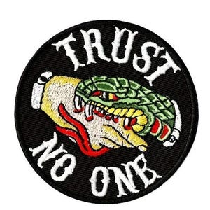Può includere: Toppa ricamata con sfondo nero. La toppa presenta una mano morsa da un serpente verde. Le parole "TRUST NO ONE" sono ricamate in bianco attorno al bordo della toppa.