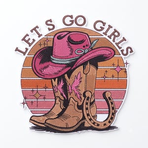 Könnte beinhalten: Ein bestickter Aufnäher mit einem rosa Cowboyhut auf einem beigen Cowboystiefel mit rosa Flügelakzenten und einem Hufeisen. Der Hintergrund zeigt einen Sonnenuntergang mit orangefarbenen und rosa Streifen sowie den Text LET'S GO GIRLS in braun.