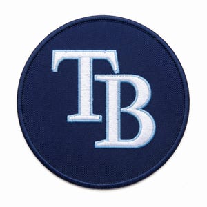 Può includere: Una toppa ricamata circolare con uno sfondo blu scuro e le lettere TB in bianco, delineate in azzurro. Le lettere sono stilizzate con un carattere serif e sono centrate sulla toppa, che presenta un bordo cucito di colore blu scuro.