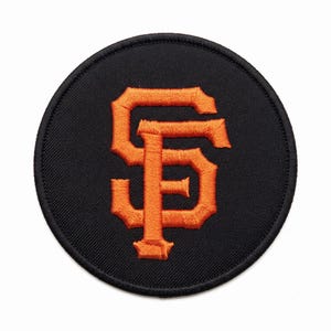 May include: A circular embroidered patch featuring a black background with a bold, orange, stylized interlocking SF logo in the center. The patch has a thick, black stitched border around the entire circumference, creating a clean, finished edge.