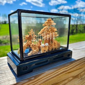 Può includere: Un diorama in miniatura in legno di un paesaggio sereno, racchiuso in una scatola di vetro con cornice nera. La scena comprende alberi in miniatura dettagliati, strutture e figurine di uccelli bianchi, il tutto su una base nera.